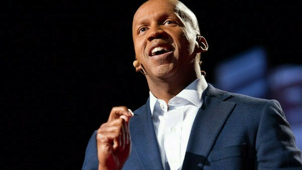 Bryan Stevenson stands in front of a dark background. His hand is raised and he is speaking. He wears a blue suit jacket and white shirt.