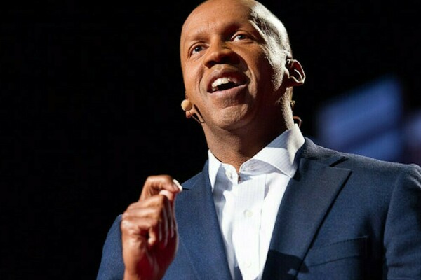 Bryan Stevenson stands in front of a dark background. His hand is raised and he is speaking. He wears a blue suit jacket and white shirt.