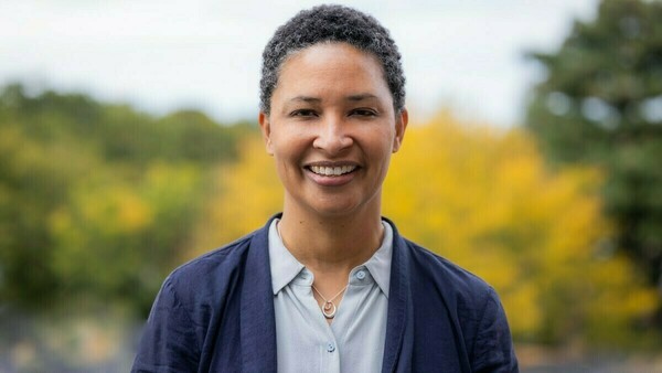 Professor Danielle Allen stands outside in front of a yellow bush. She is wearing a navy blazer over a light blue collared shirt, and she is smiling at the camera.