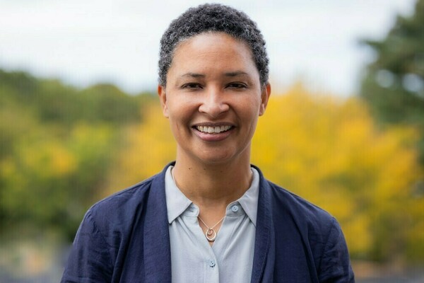 Professor Danielle Allen stands outside in front of a yellow bush. She is wearing a navy blazer over a light blue collared shirt, and she is smiling at the camera.